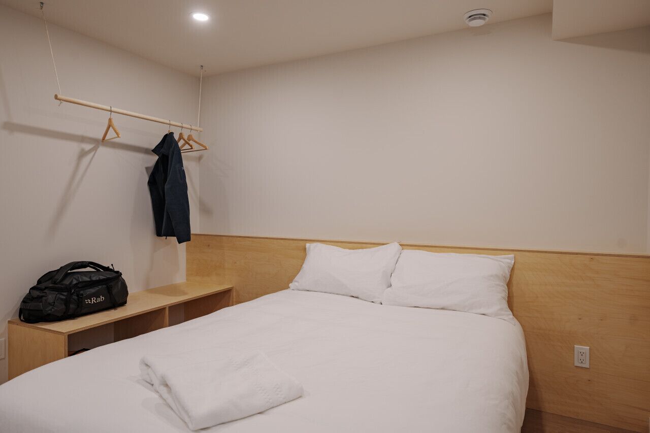 Hotellihuone, jossa on sänky, puinen pääty, yöpöytä, seinälle ripustettu naulakko ja musta laukku pöydällä.