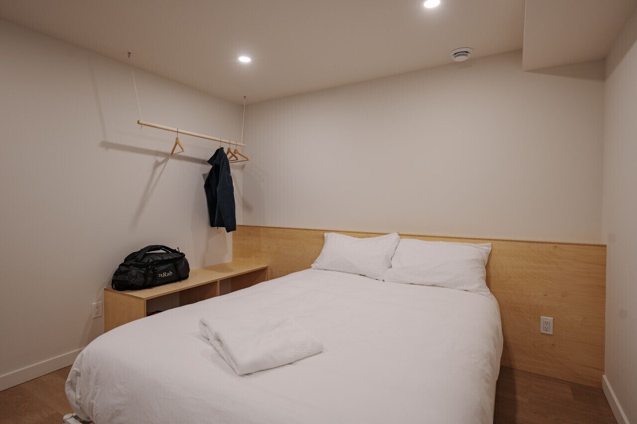 Habitación de hotel con una cama individual, cabecera de madera, mesita de noche y un perchero con ropa.