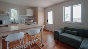 Eine moderne Küche mit Kochinsel, Barhocker und einem grauen Sofa in einem hellen Raum mit großen Fenstern.