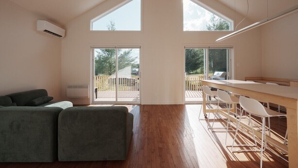 Un vaste espace de vie doté d’un haut plafond, de grandes fenêtres, d’un plancher en bois, d’un canapé vert et d’un coin bar avec des chaises blanches.