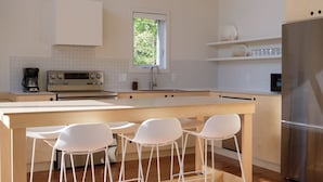 Une cuisine moderne dotée d’un îlot central, de tabourets de bar blancs, d’un réfrigérateur en acier inoxydable et d’une fenêtre laissant entrer la lumière naturelle.
