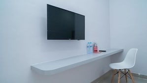 Una camera moderna con parete bianca, TV a schermo piatto montata, una sedia bianca e un piccolo ripiano con bottiglie d'acqua e un telecomando.