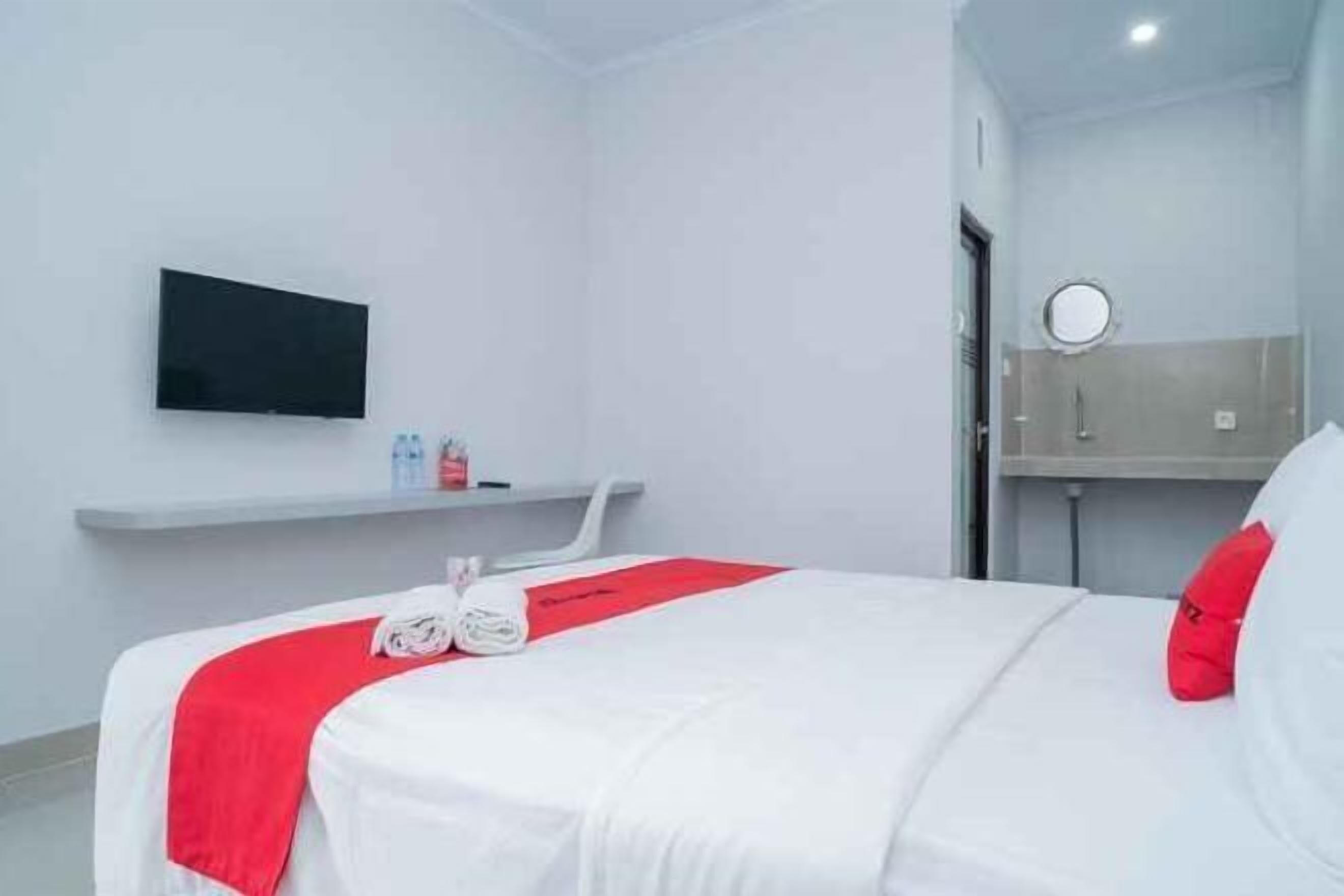 Et hotelværelse med en seng, et fjernsyn ophængt på væggen, en lille hylde med toiletartikler og et rundt spejl over vasken.