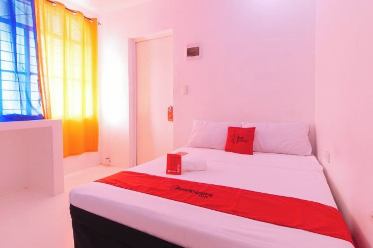 A hotel room with a bed, a red throw blanket, and a window with yellow curtains.