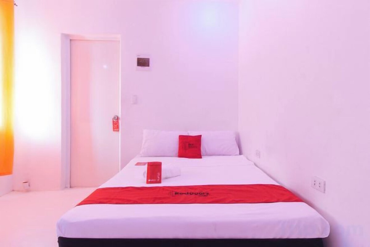 A single bed with a red blanket and pillows, a white wall, and a door to the left.