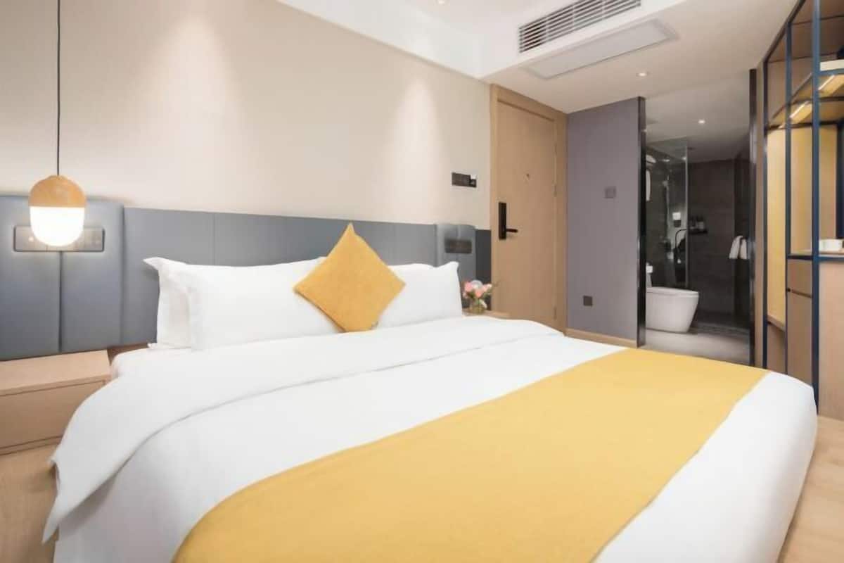 Une chambre d’hôtel moderne avec un grand lit, une table de chevet, une lampe fixée au mur et une salle de bain visible à travers une porte ouverte.