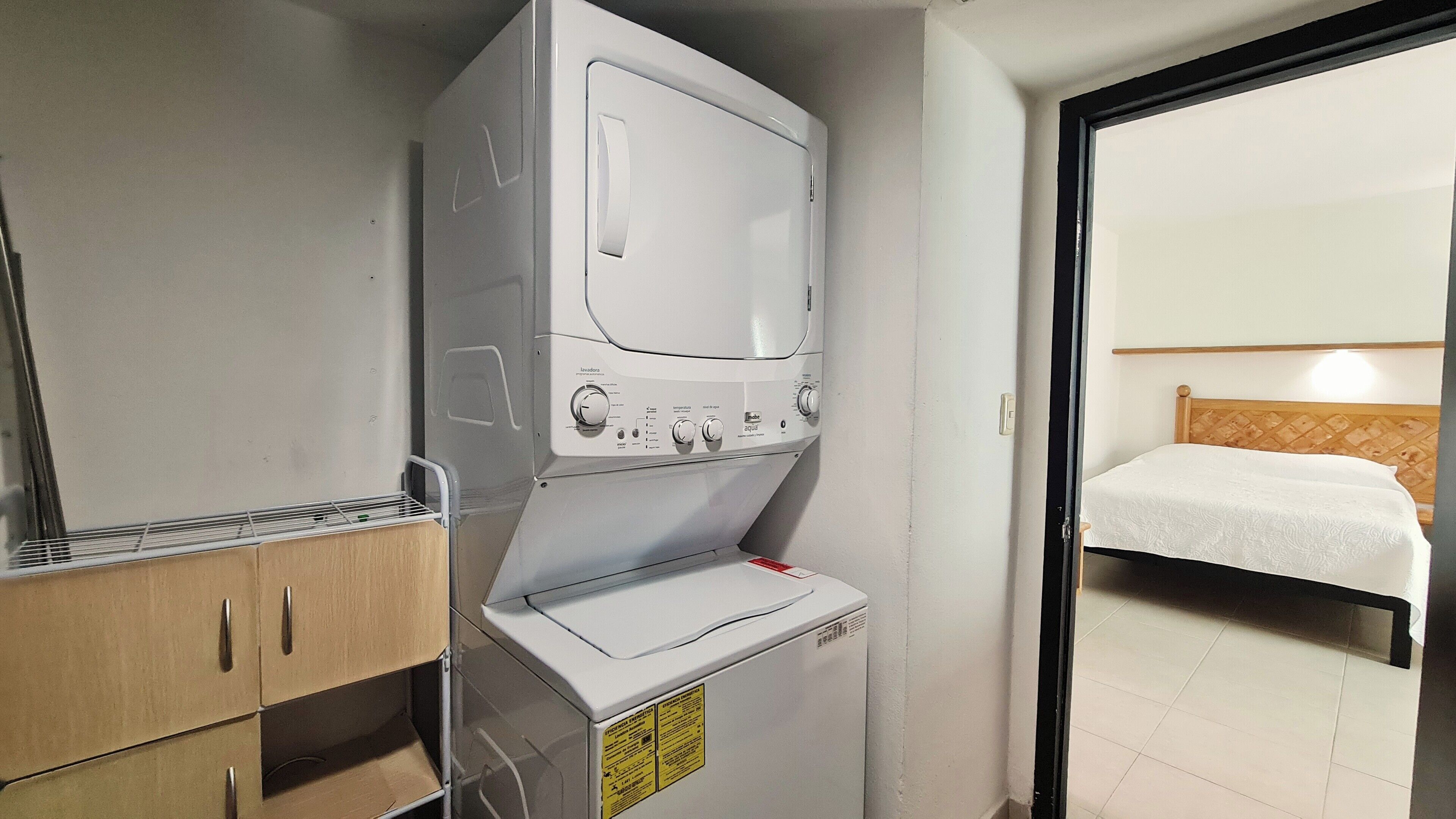 Een compacte hotelkamer met een wasmachine, een klein kastje en een bed met houten hoofdeinde.