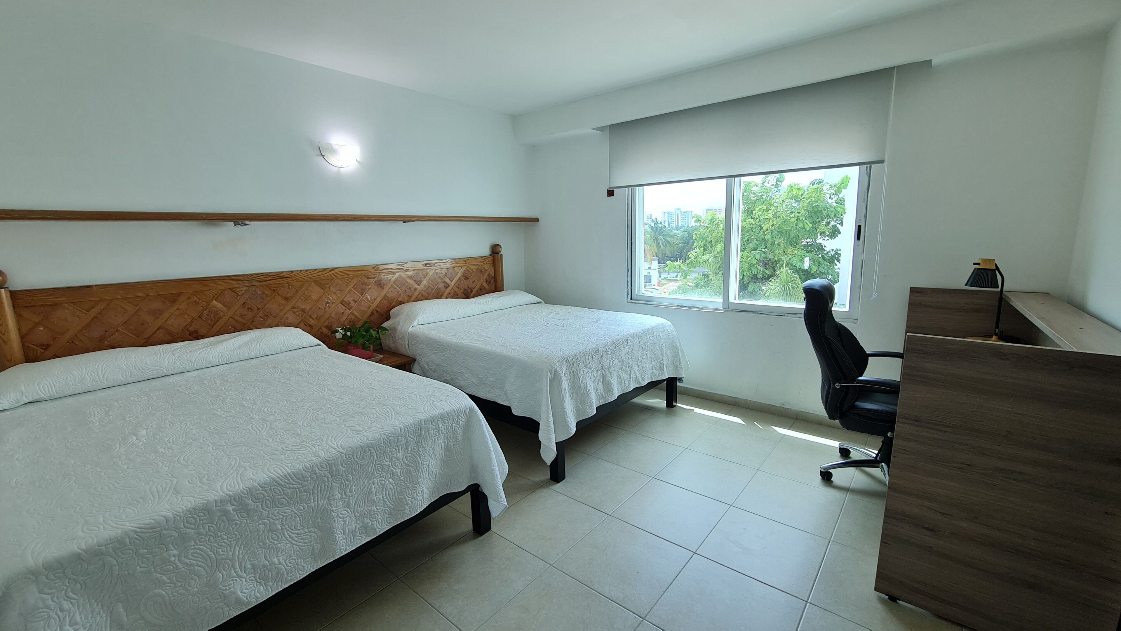 Une chambre d’hôtel avec deux lits, un bureau, une chaise, une fenêtre donnant sur un espace verdoyant et un luminaire au plafond.