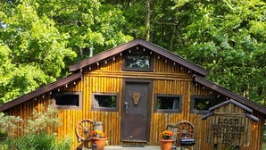Una cabaña de madera rústica con puerta principal, dos ventanas y un letrero que dice "Lost Mansions Mining Co." frente a un exuberante fondo verde.