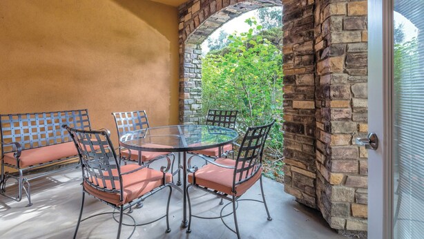A patio with a glass table and metal chairs, surrounded by stone walls and a view of greenery.