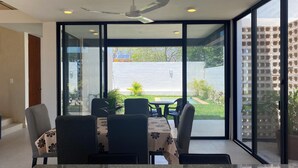 A dining area with a table and chairs, a ceiling fan, and large windows offering a view of a garden and a brick wall. - VILLA GRACE tu mejor opcion en Cholul (Mérida)
