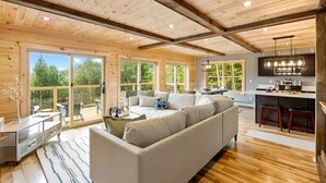 A spacious living area with a large sofa, a dining table, and a kitchen with bar stools. - Secluded Cabin with Hot Tub, Views, & EV Charger (Bryant Pond)