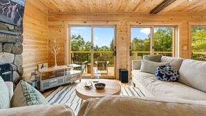 A cozy wooden cabin interior with a stone fireplace, a sofa, a dining area, and large windows offering a view of the outdoors. - Secluded Cabin with Hot Tub, Views, & EV Charger (Bryant Pond)