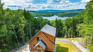 A cabin with a stone foundation and a wooden exterior, surrounded by dense forest and a lake in the background. - Secluded Cabin with Hot Tub, Views, & EV Charger (Bryant Pond)