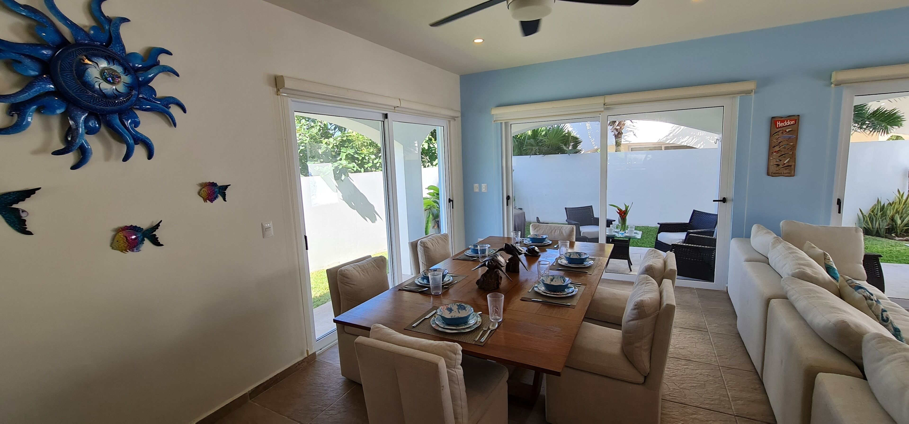 A dining area with a large table set for four, surrounded by beige chairs. There is a blue decorative wall piece above the table and a small fish decoration below it.