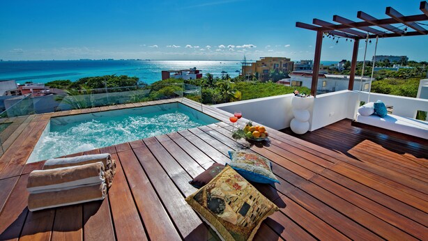 A wooden deck with a hot tub, a wooden table with towels and a fruit bowl, and a view of the ocean and buildings. - CASA REYES Stunning Rooftop Oceanview (Isla Mujeres)