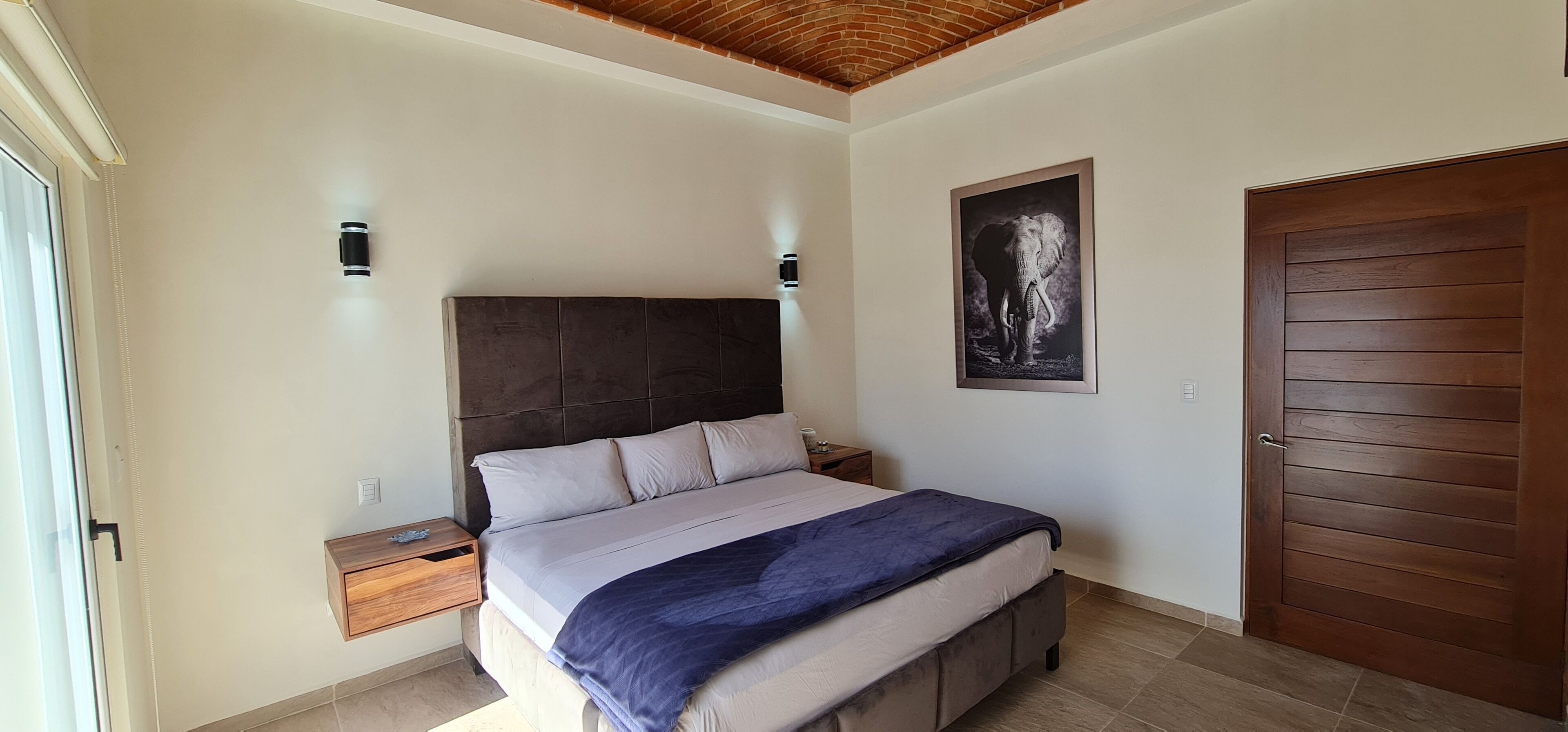 Quarto com uma cama grande, cabeceira de madeira, mesa de cabeceira, quadro emoldurado na parede e porta de madeira.