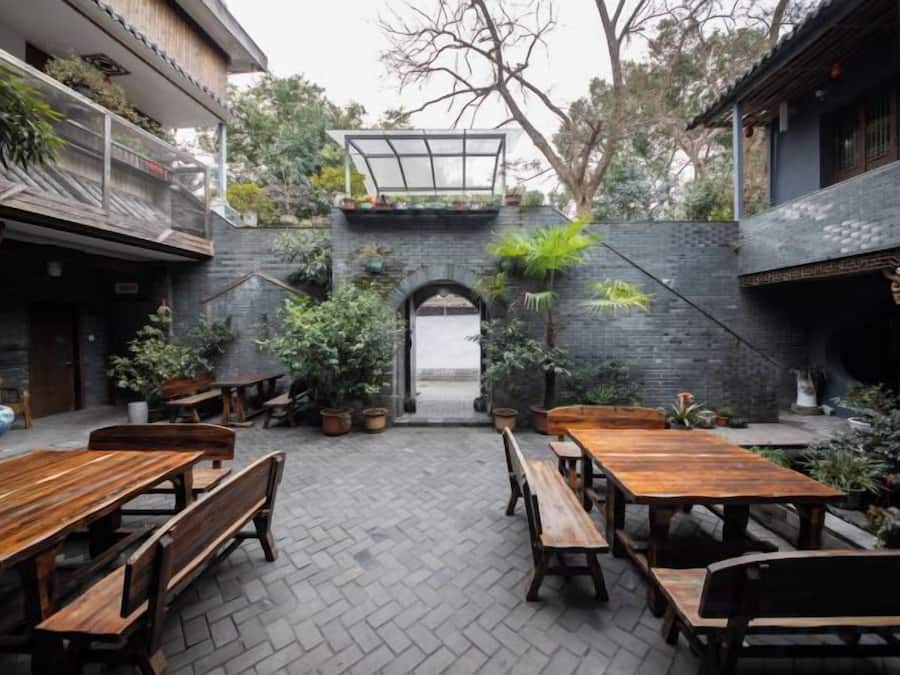 Un patio al aire libre con mesas y bancos de madera, rodeado de plantas en macetas y un muro de ladrillo.