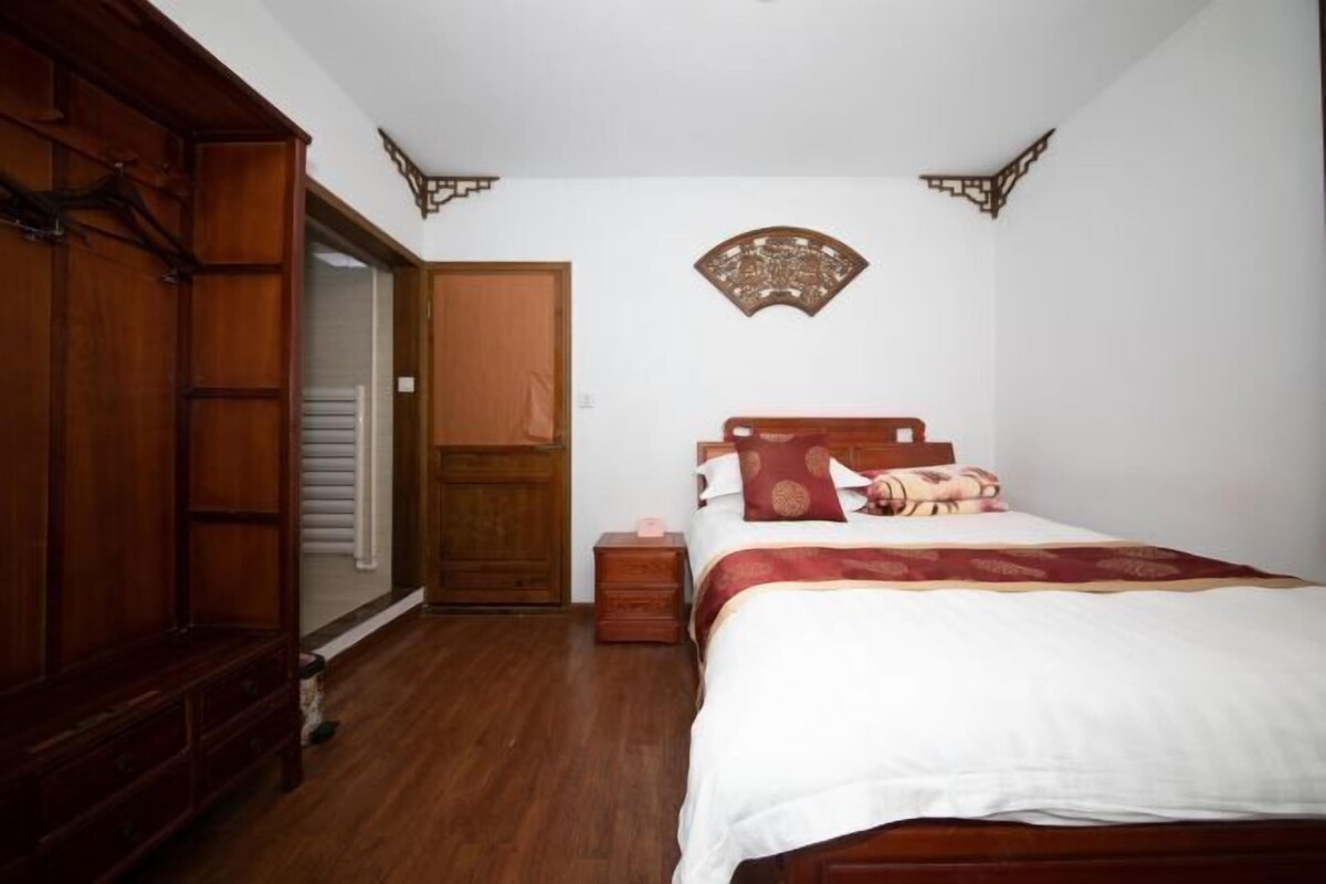 A bedroom with a wooden wardrobe, a bed with a red and white bedspread, a nightstand, and a wall-mounted decorative piece.