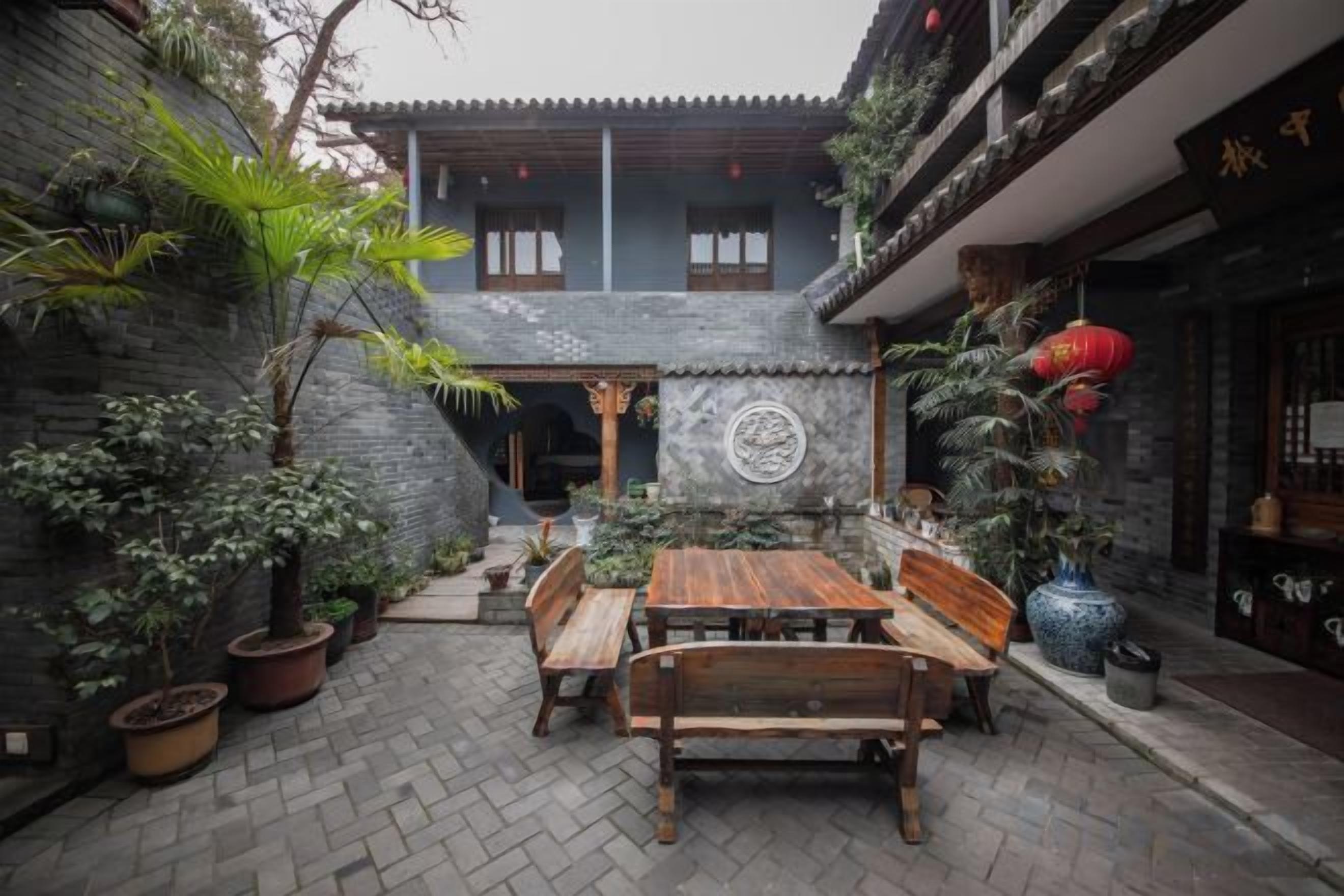 A courtyard with a wooden table and benches, surrounded by potted plants and a stone wall.