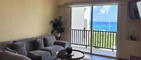 A living room with a grey sofa, a small round table, and a potted plant. There is a balcony with a view of the ocean.