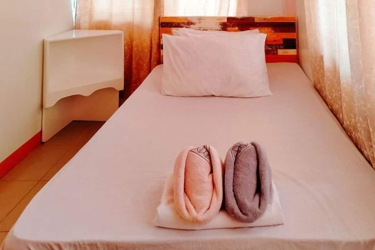 A single bed with a white pillow and two rolled towels in a room with a wooden headboard and a white nightstand.