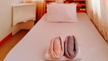 A single bed with a white pillow and two rolled towels in a room with a wooden headboard and a white nightstand.