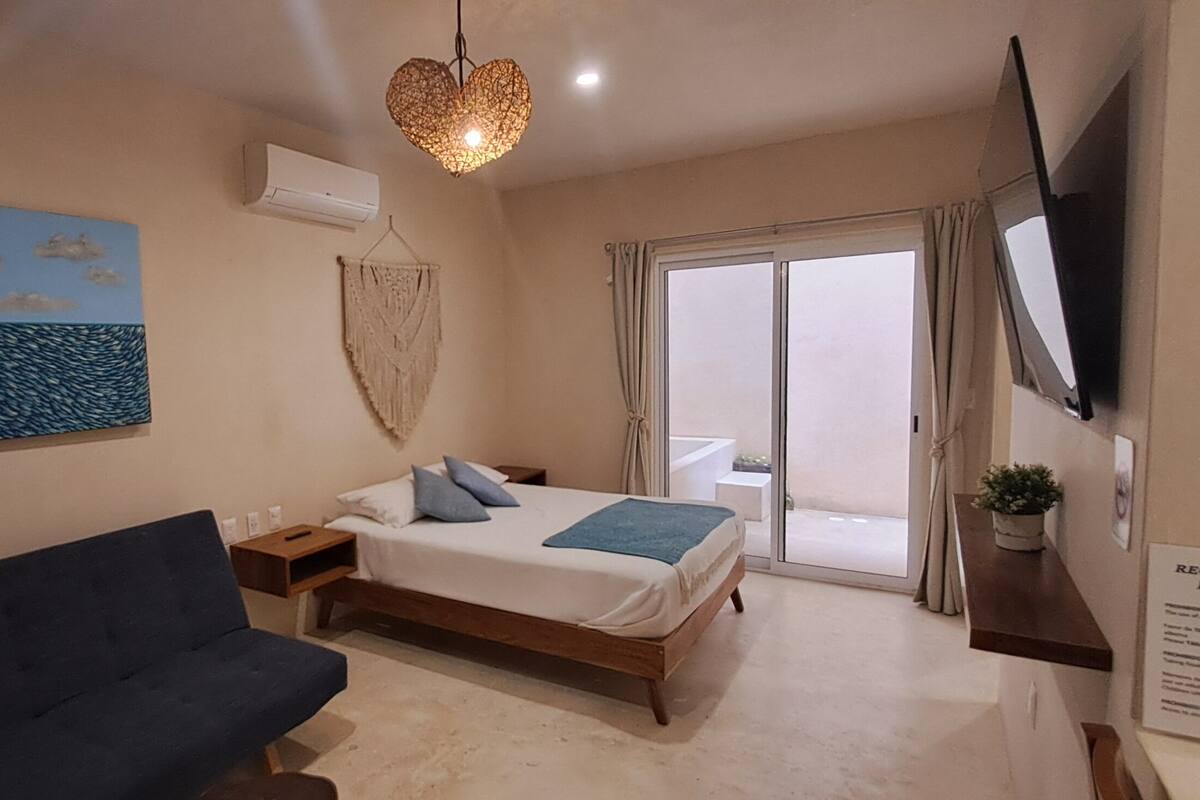 Une chambre à coucher avec un lit, un canapé bleu, un tableau représentant un voilier, une décoration murale en macramé et un téléviseur fixé au mur.