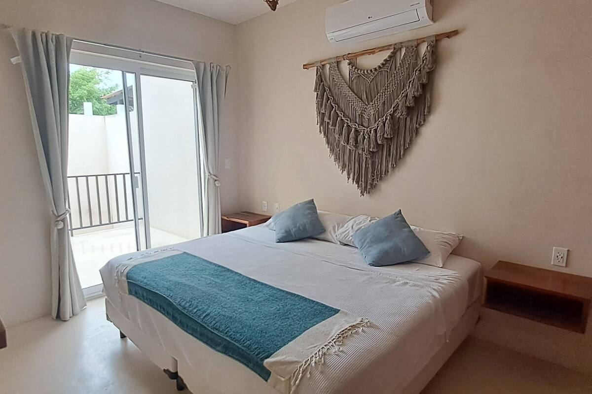 Une chambre avec un lit, une télévision, une décoration murale en macramé et un balcon avec des rideaux.