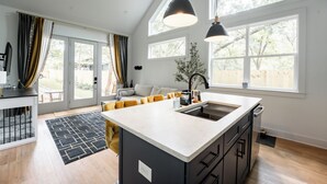A modern kitchen with a large island, a built-in oven, and a sink. There are large windows providing natural light and a view of the outdoors. - Inner City Hideout: Serenity with Big Nature Views (Atlanta)