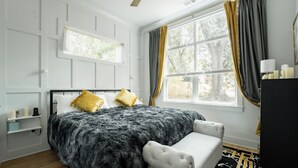 A bedroom with a bed, a white chair, a dresser, and a window with curtains. - Inner City Hideout: Serenity with Big Nature Views (Atlanta)