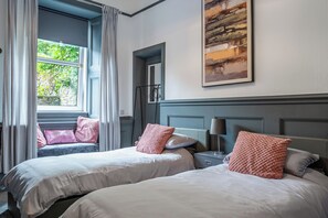 Une chambre d’hôtel avec deux lits, une grande fenêtre avec des rideaux, un tableau au mur et une table de chevet avec une lampe.