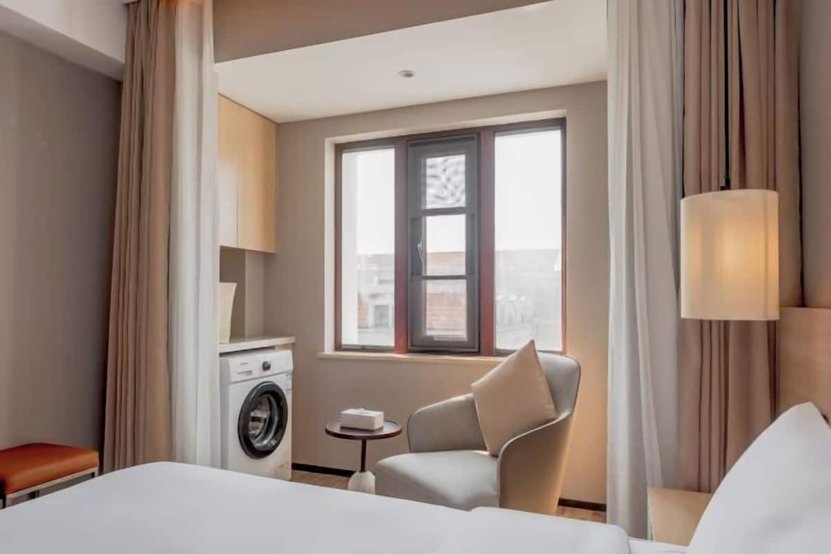 A hotel room with a bed, a chair, a washing machine, a small table, and a window with a view of buildings.