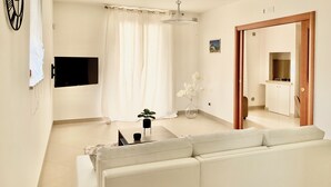 A modern living room with a white sofa, a flat-screen TV mounted on the wall, a ceiling light, and a wooden door leading to a kitchen. - Luxury Apartments  (Polignano a Mare)