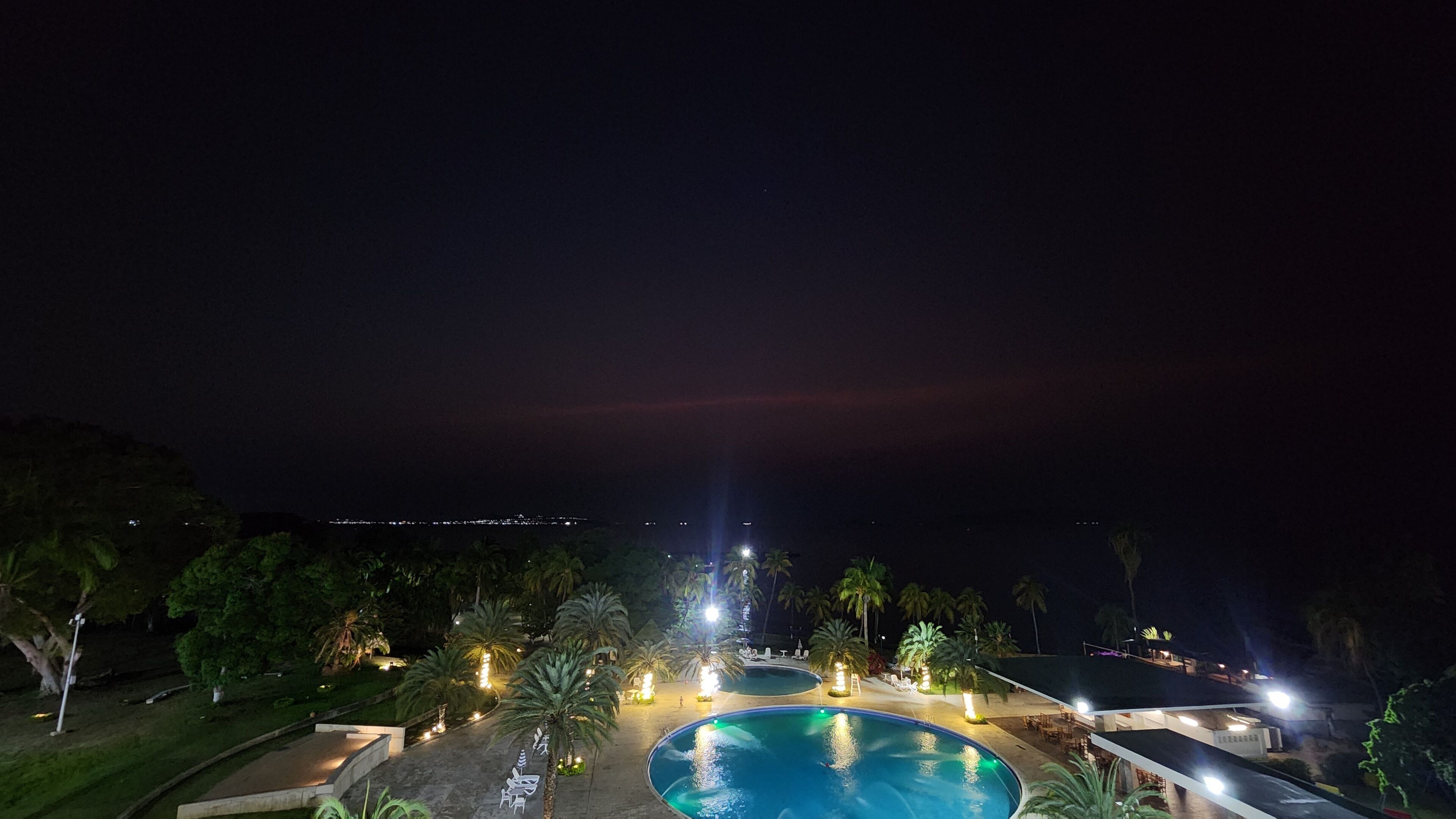 A nighttime view of a resort with a large swimming pool, palm trees, and a distant cityscape.