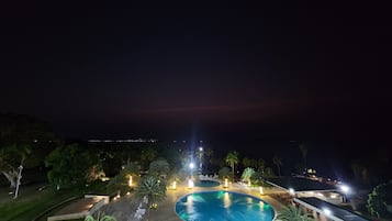 A nighttime view of a resort with a large swimming pool, palm trees, and a distant cityscape.