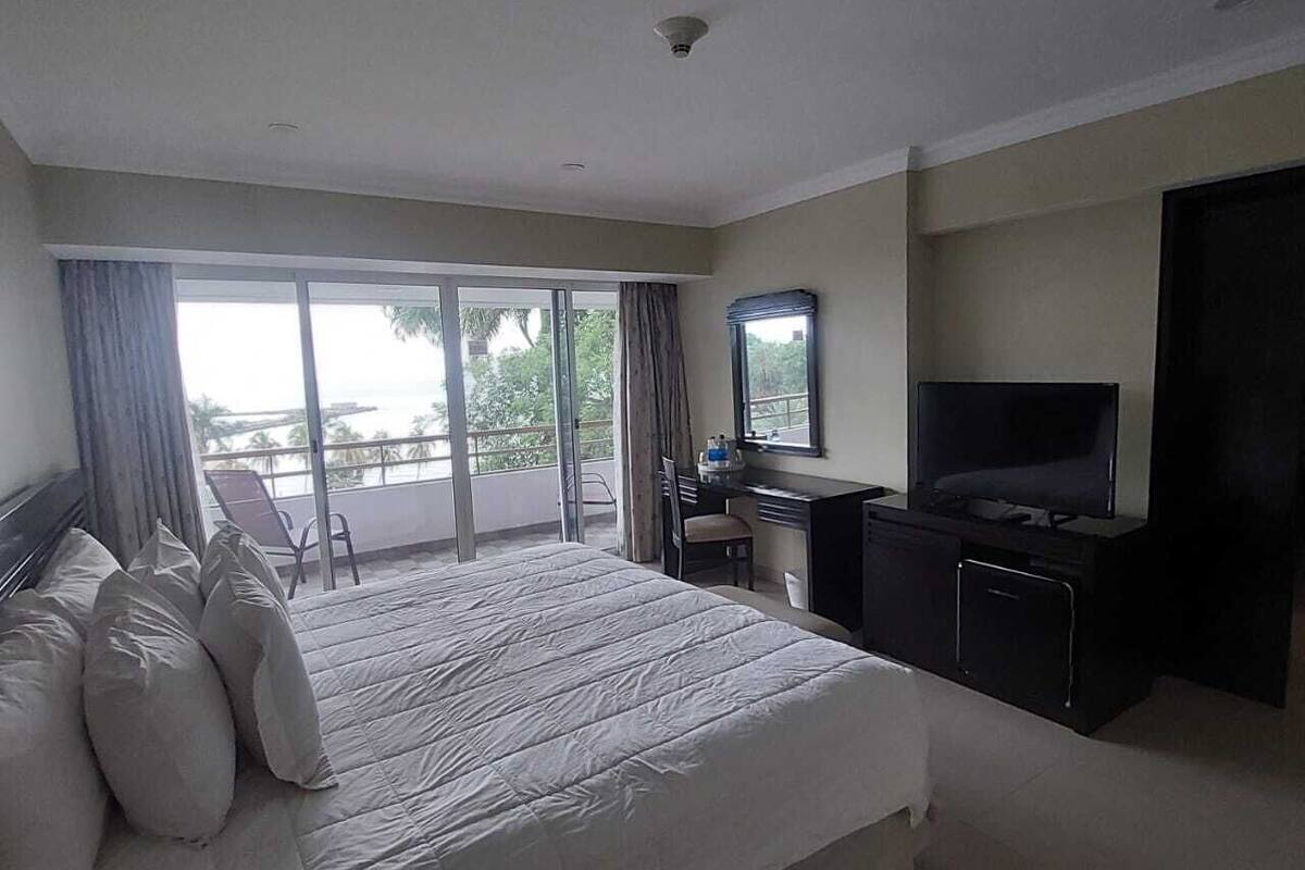 A hotel room with a large bed, a desk with a TV, and a balcony with a view.