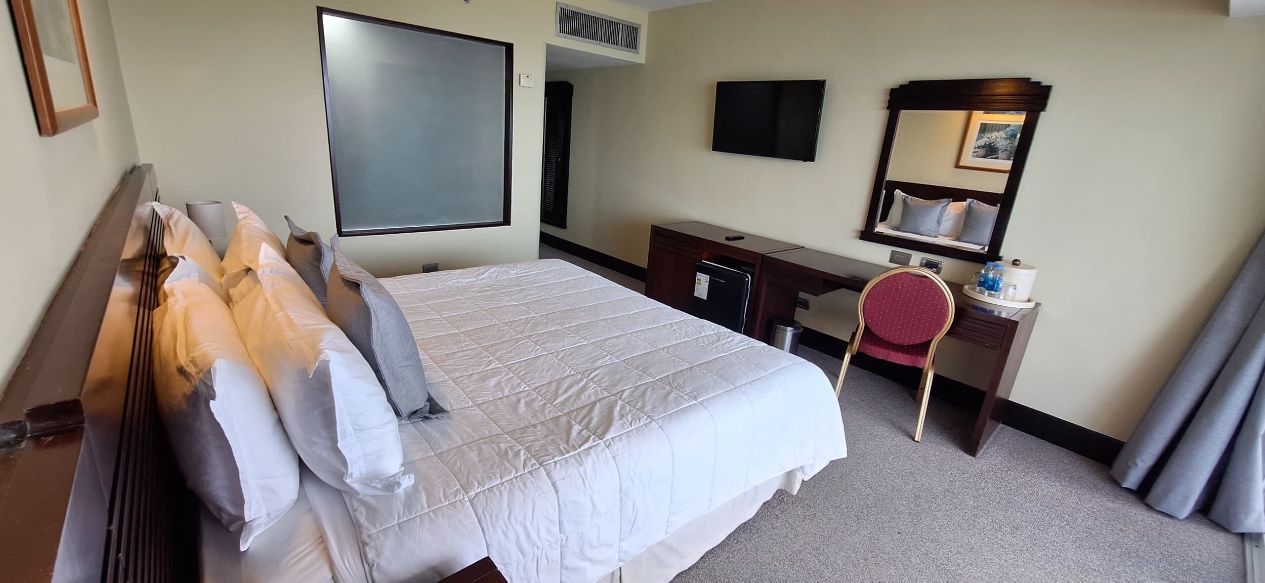 A hotel room with a large bed, a desk with a chair, a flat-screen TV, a mirror, and a small table with a cup and bottle.