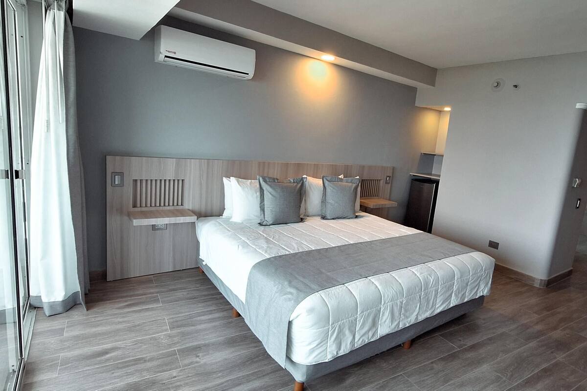 A modern hotel room with a large bed, built-in headboard, and a sliding door leading to a balcony.