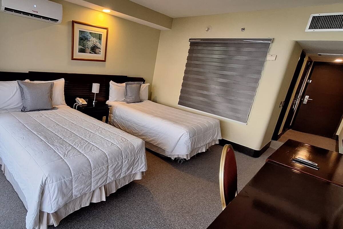 A hotel room with two beds, a desk, a chair, an air conditioner, a picture on the wall, and a window with blinds.