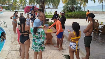 A group of people gathered around a table on a beach with palm trees and a pool in the background.
