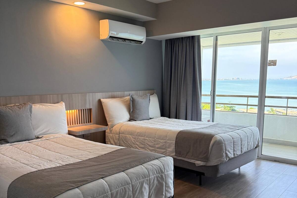 A modern hotel room with two beds, a large window with a view of the sea, and an air conditioning unit on the wall.