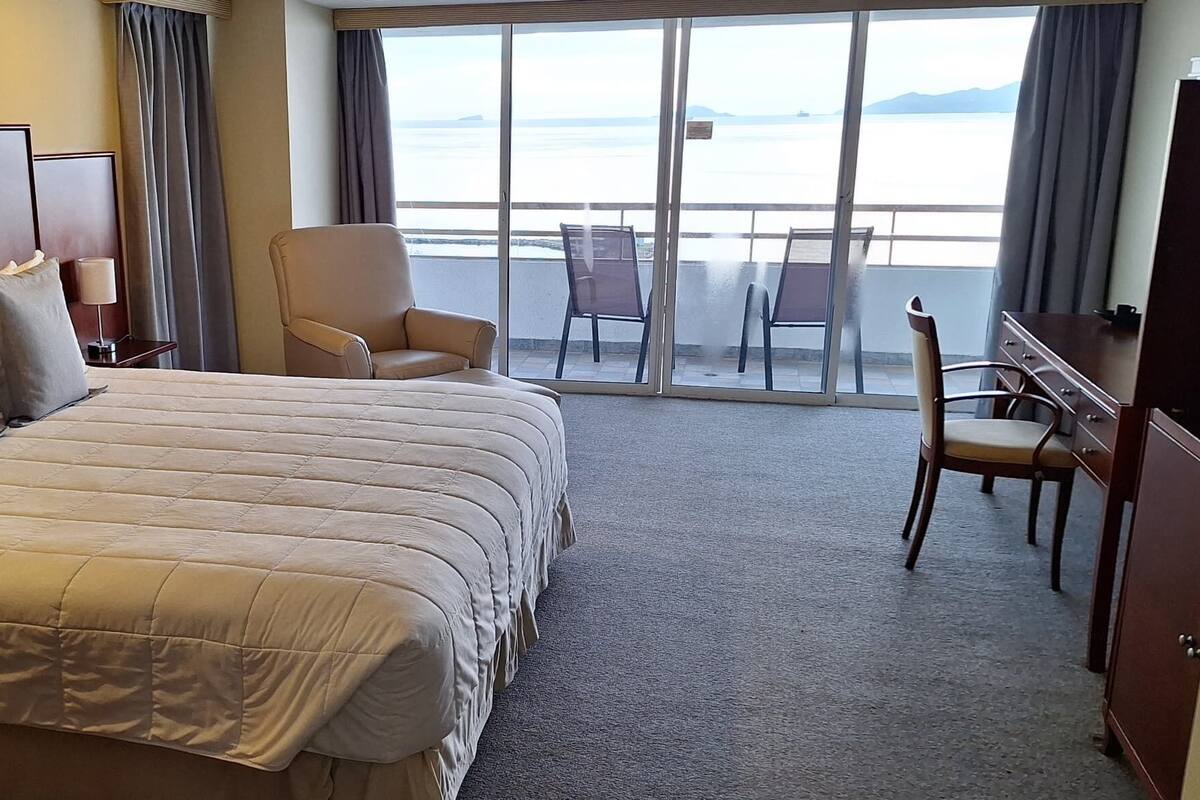 A hotel room with a large bed, a desk, and a chair. There is a view of the sea through the windows.