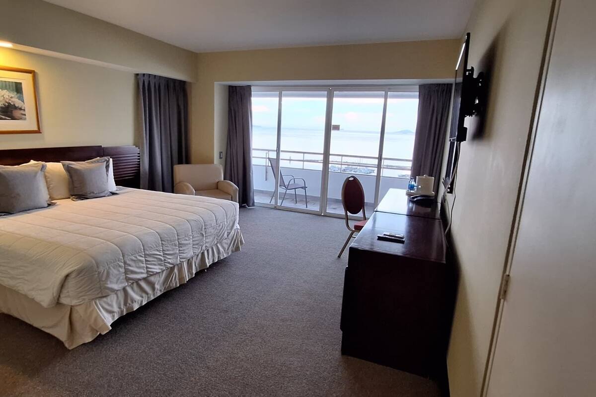 A hotel room with a large bed, a desk, a chair, and a view of the sea through a sliding glass door.