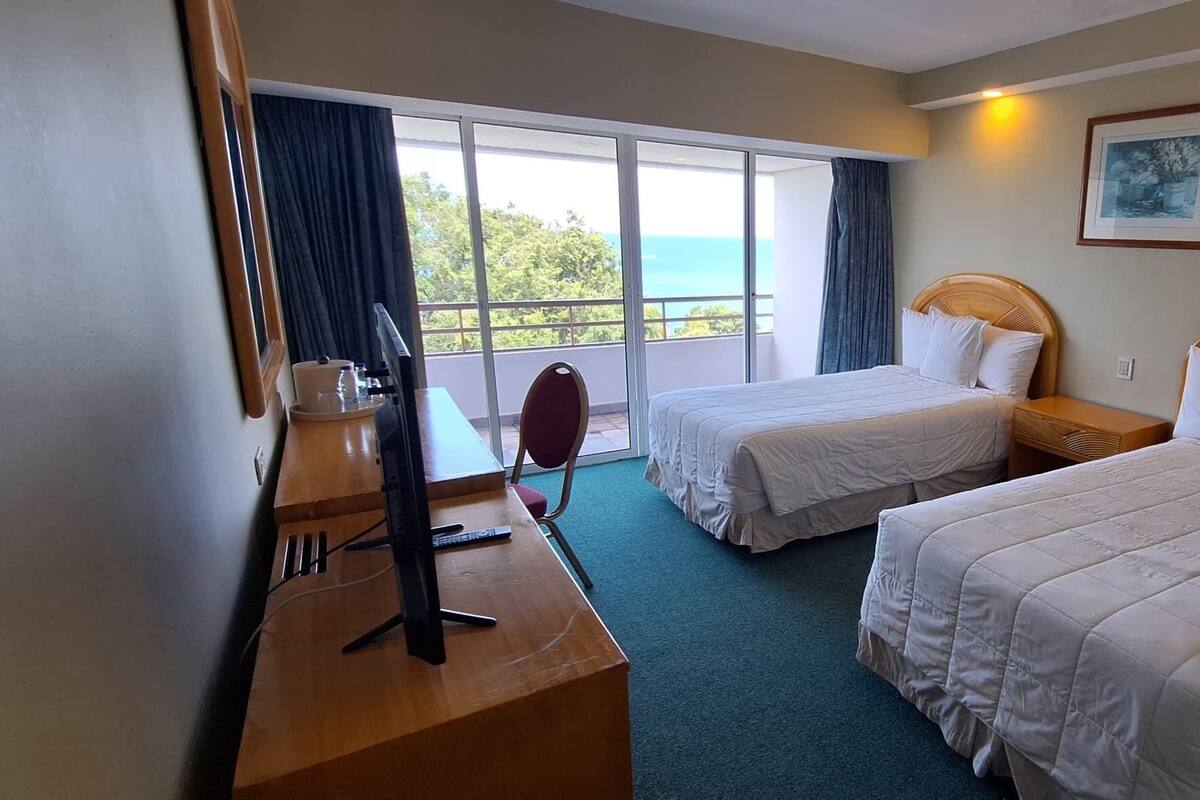 A hotel room with two beds, a desk, a chair, and a large window with a view of the sea.