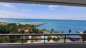 Vista desde un balcón sobre una zona costera con cielo despejado, un puerto deportivo y abundante vegetación.