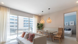 Een moderne woonkamer met een bank, eettafel en stoelen. Er is een groot raam met gordijnen, een plant en uitzicht op gebouwen.