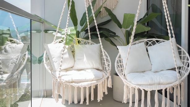 Twee macramé hangstoelen met witte kussens, een macramé wandkleed en grote groene planten.