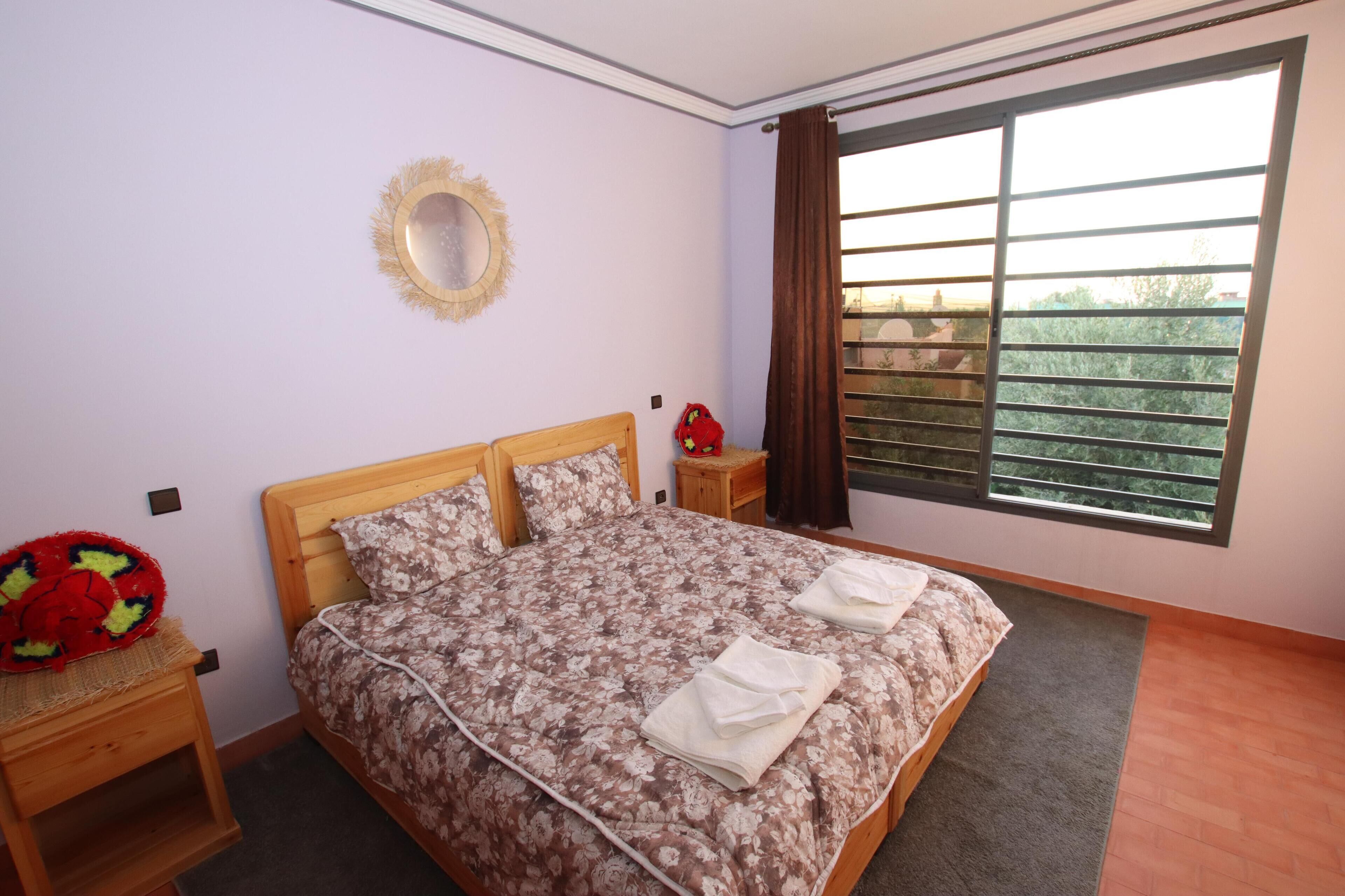 Un dormitorio con cama, mesita de noche, espejo y ventana con cortinas.