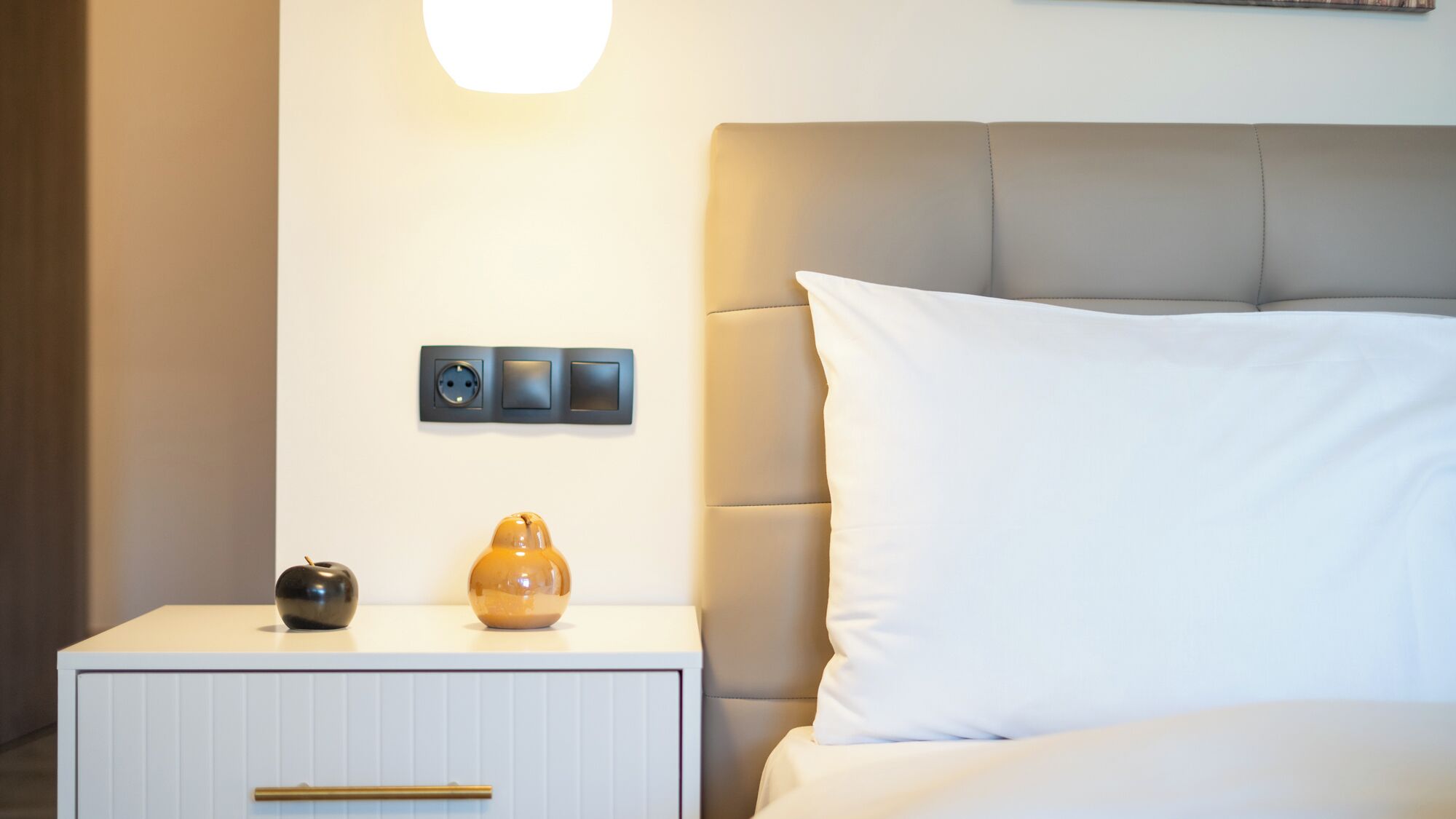 A neatly made bed with a white pillow and a beige headboard. A white nightstand with a drawer and two small decorative items is visible.